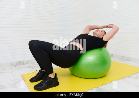 Young sporty female athlete performing exercises with a fitness ball at home studio. Engaged in a workout, leaning back on a green exercise ball. Fitn Stock Photo