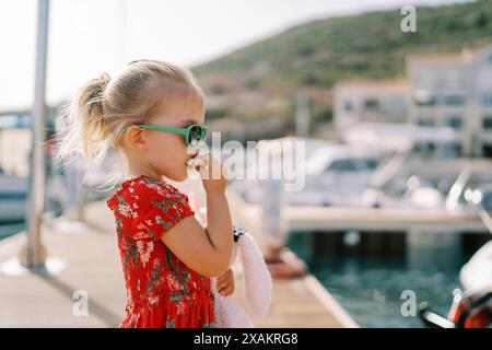 Little girl eats a bagel while standing with a toy rabbit on the pier. Side view Stock Photo