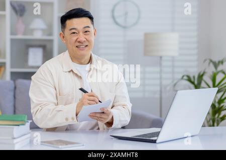 Smiling man working from home taking notes while using a laptop. Relaxed and productive atmosphere. Stock Photo