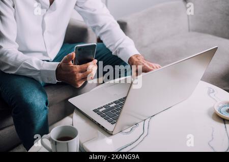 Business man in a living room, working on laptop computer and using mobile phone. Freelancer online worker searching information on internet. Networki Stock Photo