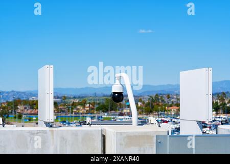 Close-up of a surveillance camera mounted on a wall, monitoring a densely packed parking lot in Los Angeles with blue sky and mountains background. Stock Photo