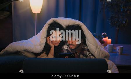 Multiracial couple man and woman shocked watching horror film movie scared afraid frightened watch video with mobile phone hiding under blanket cover Stock Photo