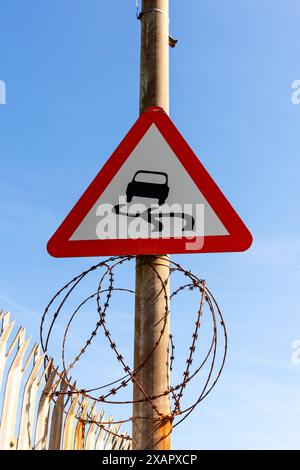 Slippery road sign on a rusty pole over some barbed wire and next to a rusty fence. The background is the bright blue sky. Stock Photo
