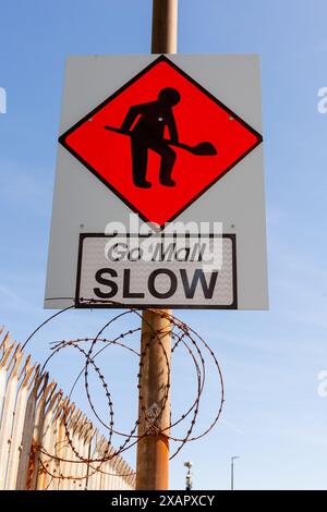 Road works and Go Slow road sign on a rusty pole over some barbed wire and next to a rusty fence. The background is the bright blue sky. Stock Photo