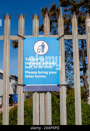 Blue, bring your litter home sign on a metal fence. There are bins, trees and grass behind the fence with a blue sky and no clouds Stock Photo