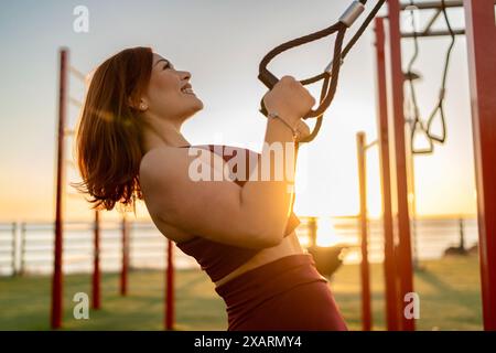 Young sportswoman is improving her fitness by doing suspension training at an outdoor gym by the sea Stock Photo