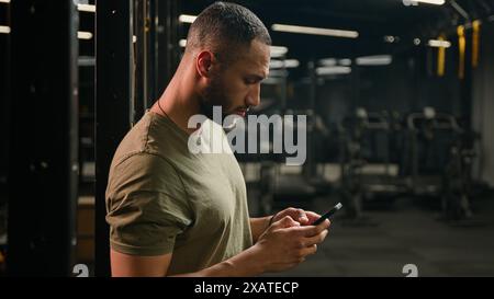 African American sportsman using mobile phone male fitness trainer man athlete relax in gym after workout exercise sport break browsing smartphone Stock Photo