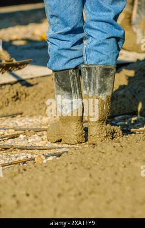 Worker's boots coated in wet concrete as it is spread over a bed of gravel and rebar at a construction site Stock Photo