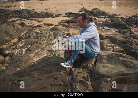 A man in casual clothes sits on a rocky beach and uses a tablet. He appears focused, perhaps working or enjoying casual browsing while surrounded by t Stock Photo