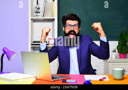 Excited teacher or student with raised hands celebrating success, victory or good result sitting at table in classroom. Bearded emotional professor in Stock Photo