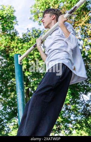 A young man in a white t-shirt and black pants is doing pull-ups on a metal bar in a park. The bar is attached to a green pole. The young man is looki Stock Photo