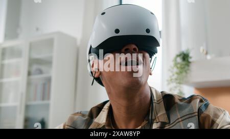 Amazed excited African guy gamer man game addict male at hone indoors in VR glasses helmet virtual reality using controllers play online 3d cyberspace Stock Photo