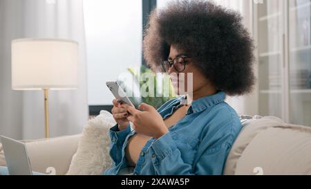 African American business woman ethnic girl freelancer student user at couch sofa at home with laptop relax work pause scrolling smartphone browsing Stock Photo