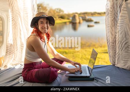 Red-haired woman teleworking typing with laptop in camper van in middle of nature near lake, connected to internet, looking at camera and smiling, Stock Photo