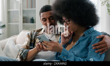 Happy married African American couple man and woman at home couch looking mobile phone shopping website app store internet using smartphone search Stock Photo