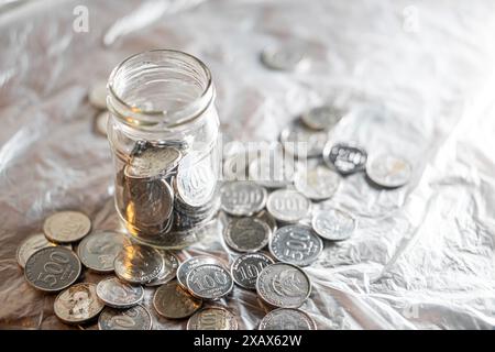 A glass jar filled with various coins, surrounded by more coins scattered on a reflective surface. Stock Photo