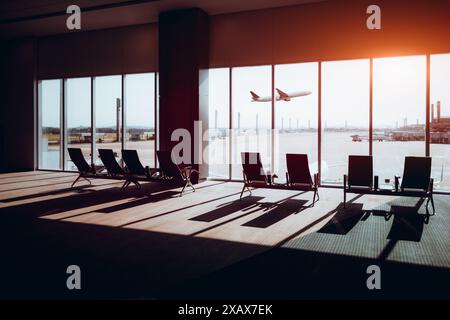 An airport modern lounge with empty seats facing large windows, overlooking the runway. A plane is taking off in the background with sunlight streamin Stock Photo