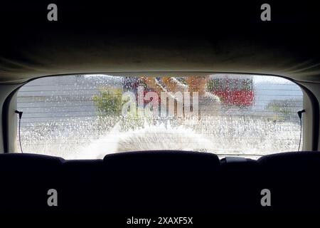 Washing the rear window of a car - view from inside the car. Close-up of glass with foam and streaks and a man with a pressure washer gun Stock Photo