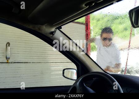 At a manual car wash: a man photographed from inside the car washes the front of a car with a high-pressure washer gun. Stock Photo