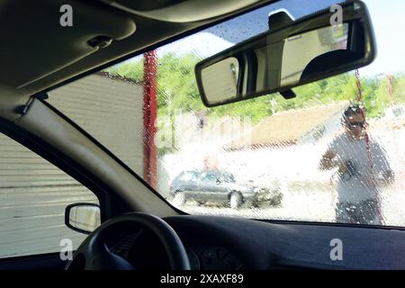 A man at a touchless manual car wash washes a car's windshield with a high-pressure washer gun. The process of washing a car, photographed from inside Stock Photo
