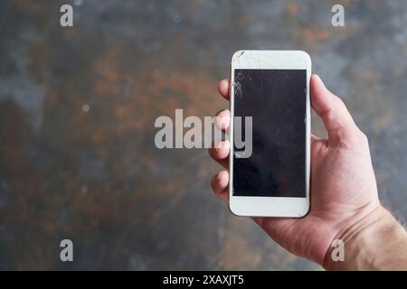 A man holds a smartphone with a broken screen in his hand on a dark background. Stock Photo