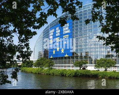 Strasbourg, France - Jun 6, 2024: The European Parliament building in Strasbourg, framed by leafy branches, featuring a large banner promoting the 2024 EU elections, with reflections on the glass facade and a river in the foreground Stock Photo