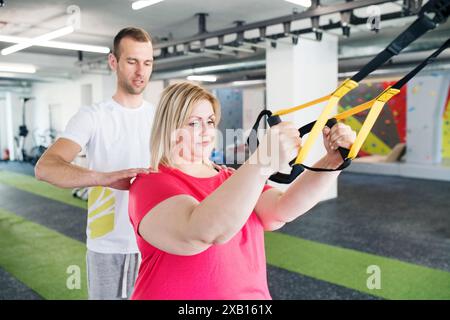 Overweight woman exercising in gym, doing suspension training, total resistance exercise. Personal trainer couching her and helping her Stock Photo