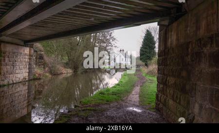 Standing on the towpath looking down the canal at a white lock keepers cottage. The image is natural framed with an old stone bridge Stock Photo