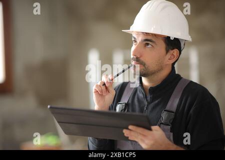Pensive construction worker holding tablet looking above in a house Stock Photo