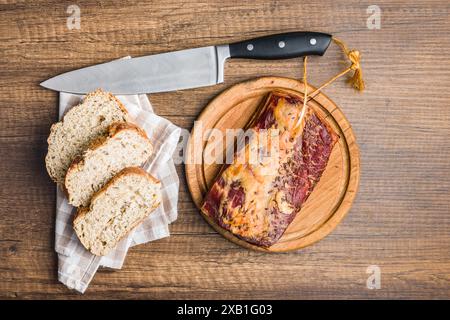 A top-down view of a wooden cutting board with a piece of caraway-spiced bacon on it, beside a stack of sliced bread on a checkered napkin. A knife li Stock Photo