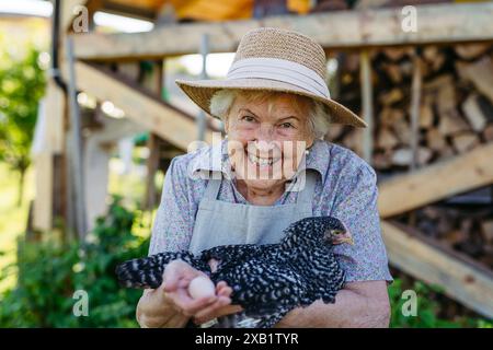 Elderly woman holding a chicken and the egg it laid. Happy old farmer is delighted with her livestock. Stock Photo