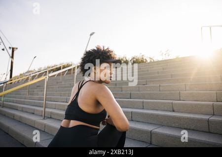 Young woman kneels on one knee, appearing to prepare for the next part of her workout. Woman warming up on steps during her exercise session. Stock Photo