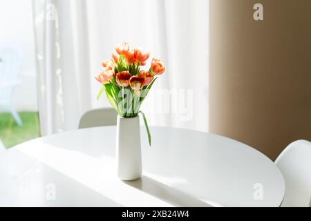 Orange tulips in a white vase on a round table, bathed in sunlight. Stock Photo