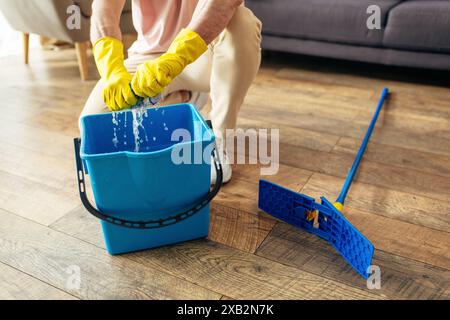 A handsome man in cozy homewear meticulously cleans a blue bucket with yellow gloves. Stock Photo
