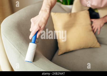 A man in cozy homewear meticulously cleans a couch using a blue sticky roller. Stock Photo