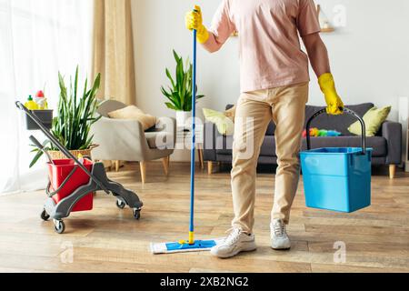 A handsome man in cozy homewear meticulously mopping floors with a bucket of cleaning supplies. Stock Photo