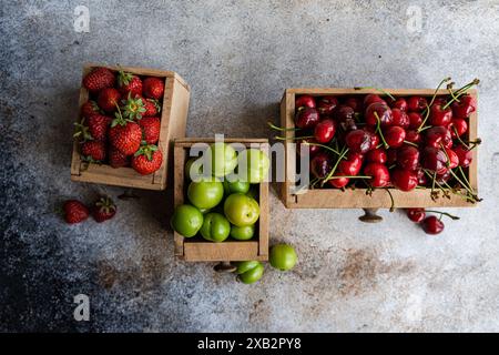 Three vintage wooden boxes showcase a selection of fresh organic berries—green plums, sweet cherries, and strawberries—on a rustic grey surface Stock Photo