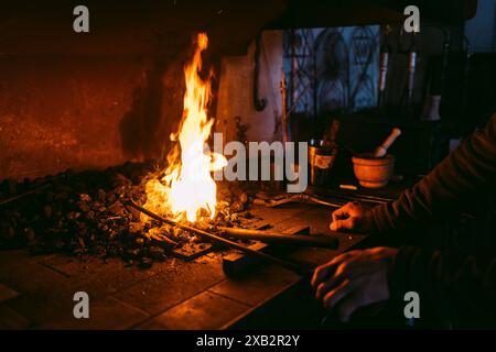 An artisan blacksmith shapes metal on an anvil with a fiery forge in the background, illuminating a dark workshop. Stock Photo