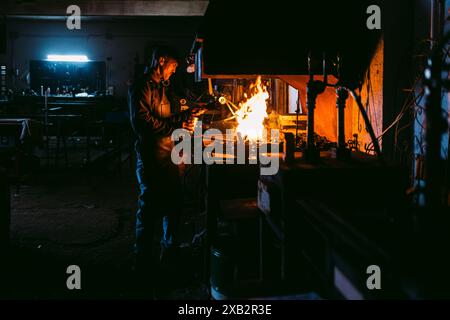 A male blacksmith is engaged in his craft, focused on heating metal in the glowing forge of a dimly lit traditional workshop. Stock Photo