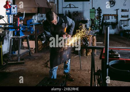 A male blacksmith wearing protective gear grinds a metal piece, with sparks flying in a dim workshop. Stock Photo