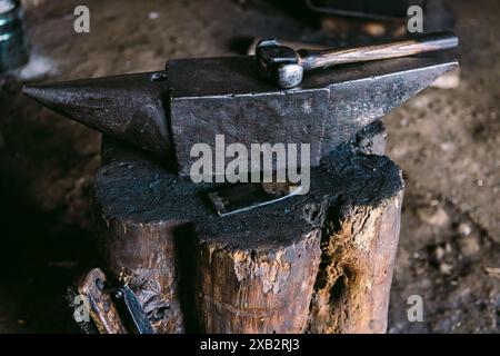 Close-up of a blacksmith's hammer and anvil set upon a sturdy wooden stump inside a rustic workshop, capturing the essence of metalworking. Stock Photo