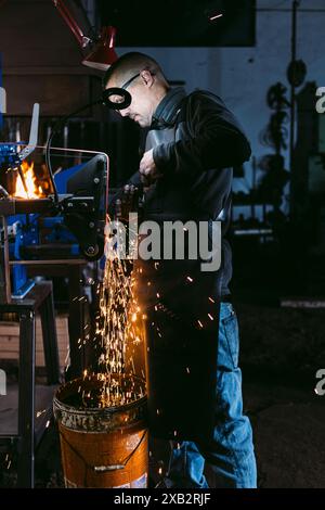A focused male blacksmith wearing safety goggles grinds metal, with sparks flying around in a dimly lit workshop. Stock Photo
