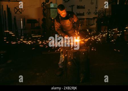 A male blacksmith is intently working on metal with a hammer over an anvil, surrounded by flying sparks in a dim workshop. Stock Photo