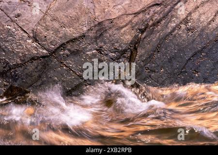 Sunlit seawater rushes against the textured surface of rugged rocks, creating a dynamic scene of natural forces at play near the sea Stock Photo