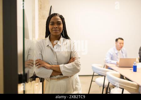 A confident woman stands with crossed arms in a modern coworking space, with team members working in the background Stock Photo