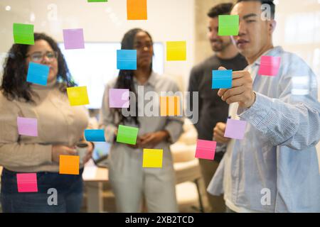 A multicultural group of friends collaborates with colorful sticky notes on a glass wall in a coworking space Stock Photo
