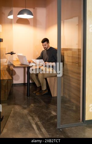 A focused man reviews documents alone in a contemporary, well-lit coworking space, embodying efficiency and concentration Stock Photo