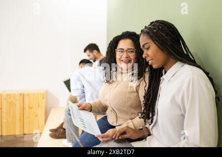 Two friends smiling and discussing work over documents in a busy coworking environment, with colleagues in the background Stock Photo