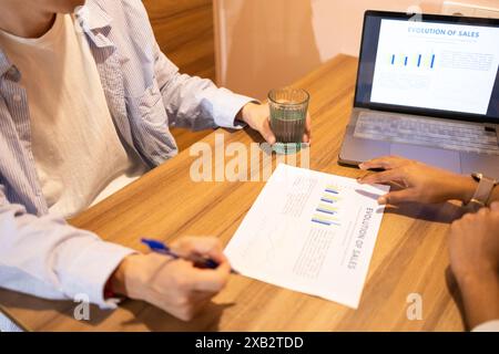 Two friends or team members are engaged in a business discussion, analyzing sales data charts on paper and a laptop in a cozy coworking environment Stock Photo