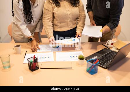 A diverse group of colleagues work together at a coworking table, discussing charts and reports, with a laptop and office supplies present Stock Photo